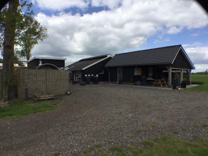 Boerderijachtig zwart houten gebouw met terras en picknicktafels, gelegen aan een open veld onder een deels bewolkte lucht.